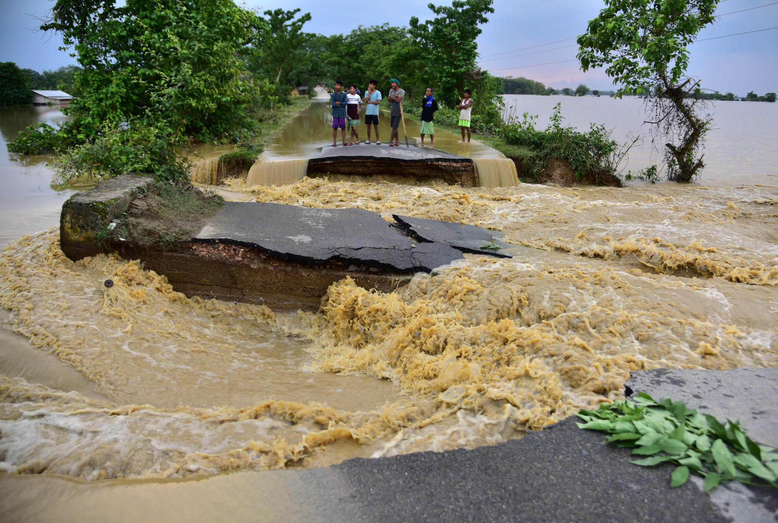 <p>People stand on a road damaged by floodwaters after heavy rains in Nagaon district, Assam, on 19 May 2022. The twin crises of extreme heat and floods are exposing a lack of preparation and shoddy infrastructure in India and Bangladesh. (Image: Anuwar Hazarika / Alamy)</p>