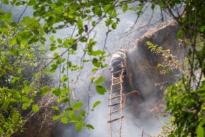 <p>Bicche Man Gurung, a honey hunter from Naiche village in Nepal’s Gandaki province, climbs down a handmade ladder to reach nests made by Himalayan giant honeybees <em>(Apis laboriosa).</em> Smoke rises from a fire lit at the bottom of the cliff by his fellow hunters to dull the bees’ senses. (Image: <a href="https://nabinbaral.com/">Nabin Baral</a> / The Third Pole)</p>
