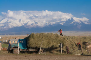 Farmers stand on the back of a truck piled with hay, in front of white snowy mountains of Kyrgyzstan