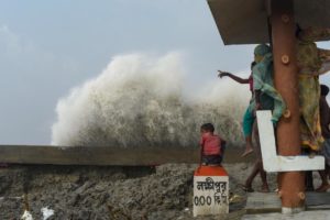 A woman and child look out to sea as a wave crashes against a tidal barrier, Cyclone Yaas. Photo by Arijit Naiya