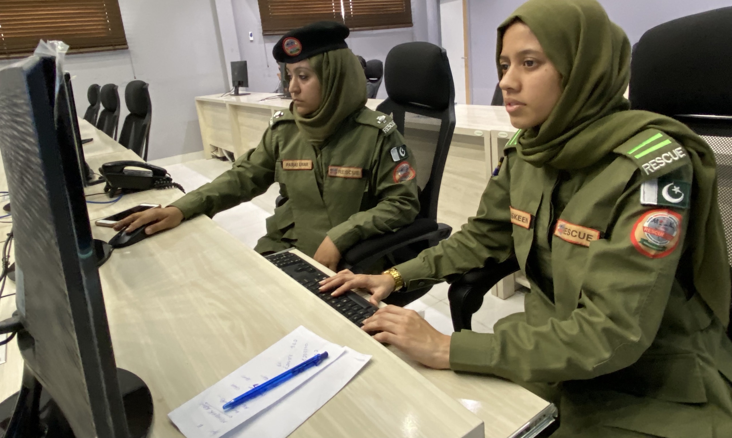 Two women in camoflague green uniform sat at a desk in front of a computer