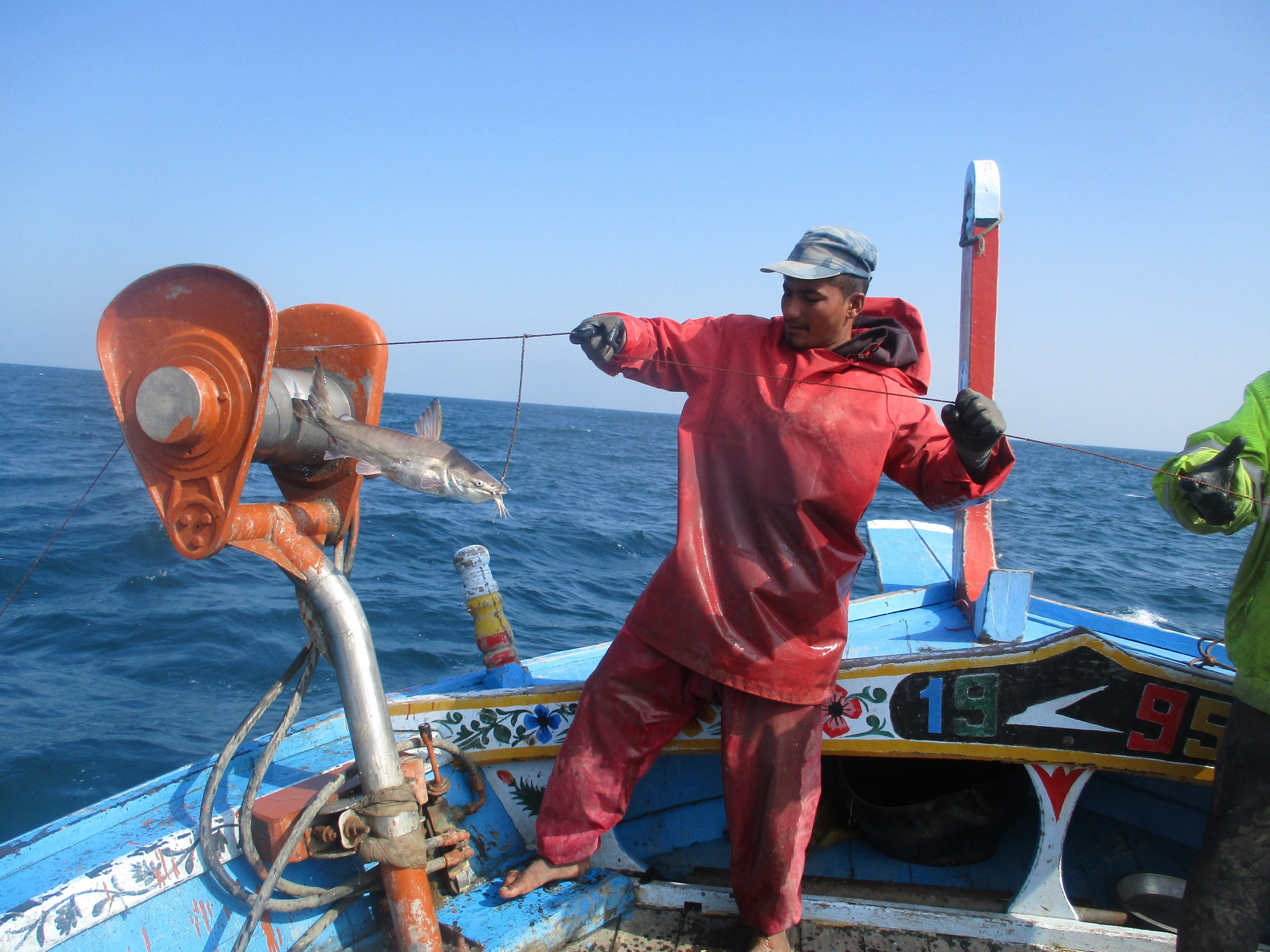 A fisherman in red overalls pulls a fishing line with a fishing hanging off, blue ocean in background