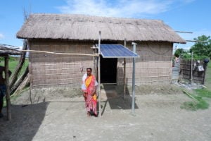 woman in pink and orange sari stands in front of a thatched house in Assam, India, with a solar panel fixed above the front door