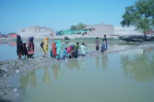 <p>Women and children crossing stagnant floodwaters in March 2023, in Dadu district of Sindh, southern Pakistan (Image: Zulfiqar Kunbhar)</p>
