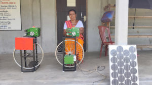 A woman sitting next to a weaving machine with a solar panel in the front