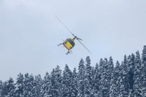 A yellow helicopter above snow-capped trees
